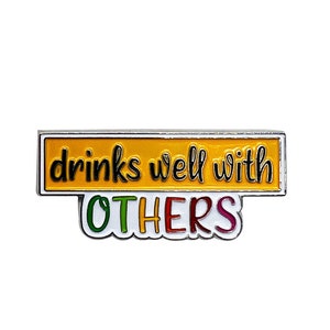 May include: A yellow enamel pin with a silver border. The pin says "drinks well with others" in black text. The word "others" is in rainbow colors.