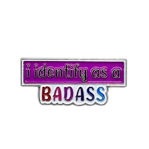 May include: A silver enamel pin with a purple rectangle that says "i identify as a" and a white rectangle below that says "BADASS" in rainbow colors.