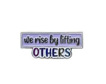 May include: A purple enamel pin with a silver border. The pin says "we rise by lifting OTHERS" in a colorful font.