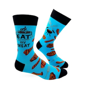 Può includere: Un paio di calzini blu con polsino nero. I calzini hanno un motivo di carne alla griglia, un barbecue e una spatola. I calzini hanno il testo "EAT my MEAT".