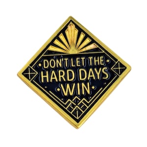 May include: A gold-coloured, diamond-shaped pin with a black interior. The pin features the text "DON'T LET THE HARD DAYS WIN" in gold lettering. The design includes art deco elements, such as sunburst and geometric patterns.