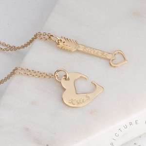 May include: Two gold necklaces with matching pendants. One pendant is a heart with the word "Kya's" engraved on it. The other pendant is an arrow with the word "Ellie" engraved on it.
