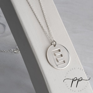 Puede incluir: Un collar de plata con un colgante redondo que presenta una letra E recortada. El colgante cuelga de una cadena de plata.
