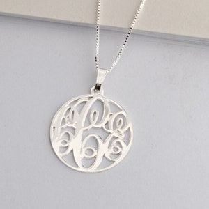 May include: A silver necklace with a round pendant featuring a monogram design. The pendant is cut out, creating a delicate and intricate look.