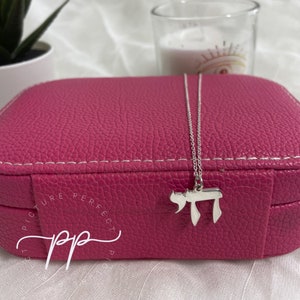 May include: A silver chain necklace with a silver charm in the shape of the Hebrew letter Chai, resting on a pink jewelry box with the words "Picture Perfect" written on it.