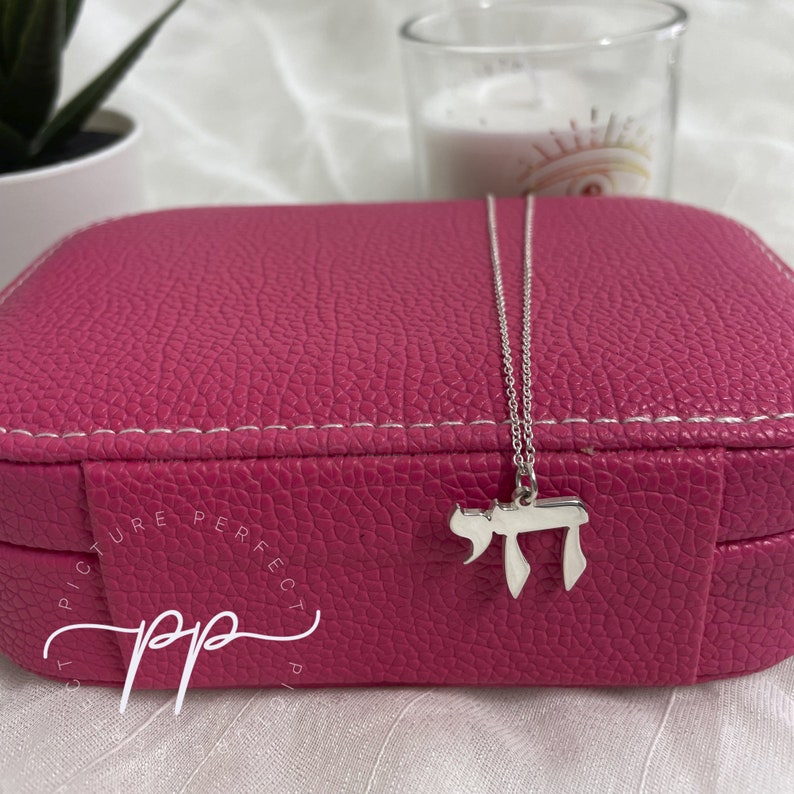May include: A silver chain necklace with a silver charm in the shape of the Hebrew letter Chai, resting on a pink jewelry box with the words "Picture Perfect" written on it.