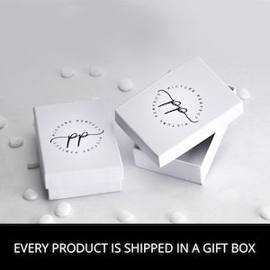 Puede incluir: Cajas de regalo blancas con el texto "PICTURE PERFECT" en un diseño circular negro. Una caja está abierta, revelando el interior. Las cajas están sobre una superficie blanca con pequeñas decoraciones blancas en forma de corazón. El texto "EVERY PRODUCT IS SHIPPED IN A GIFT BOX" está en la parte inferior.