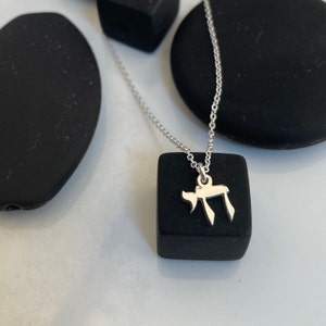 May include: A silver necklace with a small silver pendant shaped like the Hebrew letter 'Chai'. The necklace is displayed on a black square.