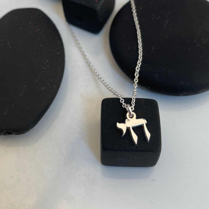 May include: A silver necklace with a small silver pendant shaped like the Hebrew letter 'Chai'. The necklace is displayed on a black square.