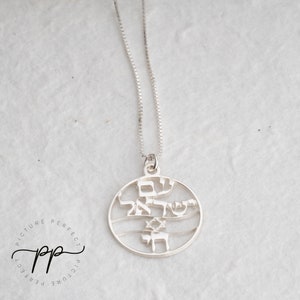 May include: A silver necklace with a round pendant featuring Hebrew text and a Star of David. The pendant is cut out, creating a delicate design.