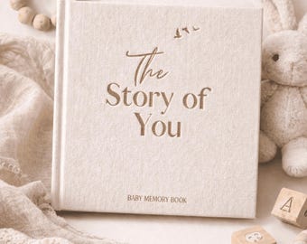 May include: A cream-colored baby memory book titled "The Story of You" with a soft fabric cover. The book is surrounded by baby items, including a stuffed bunny, wooden blocks, and a pair of knitted booties. The overall theme is baby and childhood.