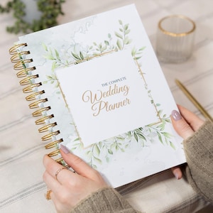 May include: A white wedding planner with a gold spiral binding and a green floral design. The cover features the text "The Complete Wedding Planner" in gold lettering.