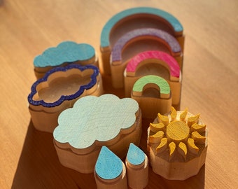 May include: A set of wooden stamps featuring various weather-themed designs, including a sun, clouds, raindrops, and rainbows. The stamps are painted in bright colors, including yellow, blue, green, and purple.