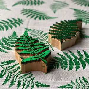 Könnte beinhalten: Zwei Holzstempel in Form von Farnblättern mit grünen Tintendetails liegen auf einem weißen Stoff mit einem grünen Farnblattmuster. Die Stempel sind rechteckig mit einem detaillierten Farnblattdesign. Der Stoff hat ein sich wiederholendes Muster aus grünen Farnblättern.