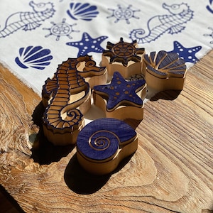Op de afbeelding: Houten stempels met blauwe inkt met zeepaardjes, zeesterren, schelpen en nautilus-ontwerpen. De stempels bevinden zich op een witte stoffen achtergrond met blauwe bedrukte zeepaardjes, zeesterren, schelpen en nautilus-ontwerpen.