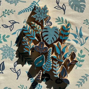 Op de afbeelding: Een set van 15 houten stempels met verschillende blad- en plantontwerpen in tinten blauw en groen. De stempels zijn op een witte stoffen achtergrond met een blauw bloemenpatroon gestapeld.