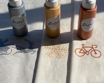 May include: Three bottles of paint in silver, gold, and copper with white lids. The bottles are labeled "EmiCraft" and have a website address. The bottles are sitting on a white fabric surface.