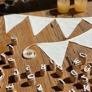 May include: Wooden alphabet stamps with white letters on a wooden surface. The stamps are arranged in a random order. There is a white fabric banner in the background.
