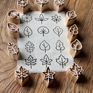 May include: A set of 18 wooden leaf-shaped stamps arranged in a circle around a piece of fabric with printed leaf designs. The stamps are made of light brown wood and have white outlines of various leaf shapes.