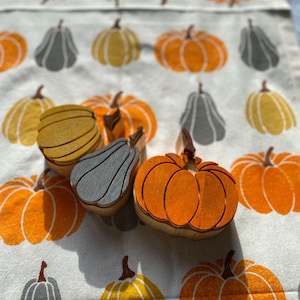 Pode incluir: Quatro carimbos de madeira em forma de abóboras e abóboras. Os carimbos estão sobre um fundo de tecido branco com um padrão de abóboras laranja e amarelas e abóboras cinzas.