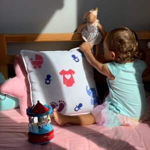 Op de afbeelding: Een wit kussen met blauwe en rode baby-thema ontwerpen, waaronder een speen, een kinderwagen, een voetafdruk, een rompertje en een olifant. Een kind zit op een roze bed met het kussen, en een houten carrousel staat op de voorgrond.