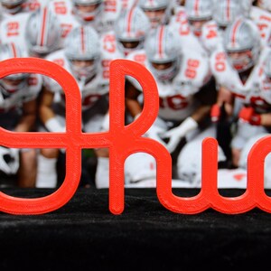 May include: A bright red, three-dimensional sign featuring the word OHIO in a stylized, connected script font. The letters are constructed with a visible layered texture, and the sign is positioned upright against a blurred background of football players.