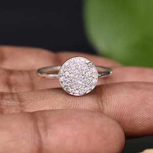 Puede incluir: Un anillo de plata con una piedra central redonda engastada en pavé. El anillo está en un dedo.