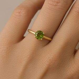 以下が含まれることがあります： 緑色の宝石がついた金の指輪。指輪は人の指にはめられています。