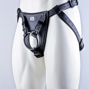 May include: Black leather harness with a metal ring and a buckle. The harness is worn on a white mannequin.