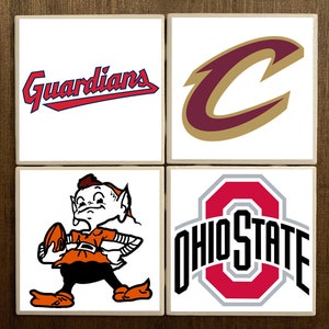 May include: Four white ceramic coasters with team logos. The top left coaster features the Cleveland Guardians logo in red and blue. The top right coaster features the Cleveland Cavaliers logo in maroon and gold. The bottom left coaster features the Cleveland Browns logo, a cartoon elf holding a football. The bottom right coaster features the Ohio State Buckeyes logo in red, black, and gray.