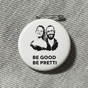 Puede incluir: Un botón blanco con una ilustración en blanco y negro de dos personas. El botón tiene el texto "BE GOOD BE PRETTI" en negro. Un imperdible plateado está sujeto en la parte posterior.