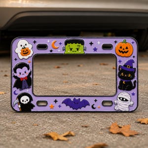 May include: A rectangular purple license plate frame featuring a variety of colorful Halloween-themed characters including a ghost, Frankenstein's monster, a pumpkin, a vampire, a black cat, a grim reaper, a bat, and a mummy with stars and moons.