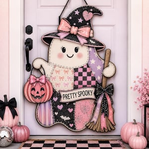 May include: A decorative wooden door hanger shaped like a ghost wearing a black witch hat with a pink bow. The ghost features a patchwork design in pink, black, and white with hearts, stars, and bats. It holds a pink pumpkin bucket and a broom.