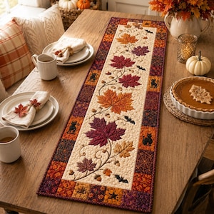 May include: A rectangular quilted table runner featuring a central cream-colored panel decorated with embroidered autumn maple leaves in shades of orange, red, and purple, interspersed with small black cat and bat silhouettes and acorns.