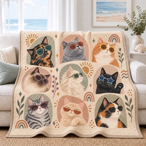 May include: A beige quilted throw blanket featuring a grid of nine illustrated cats wearing various styles of sunglasses. The cats are depicted in different colors and patterns, surrounded by small graphic icons like suns, rainbows, and leaves.