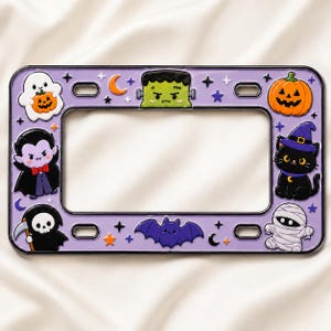 May include: A rectangular purple license plate frame featuring colorful enamel-style Halloween icons including a ghost with a pumpkin, Frankenstein's monster, a jack-o'-lantern, a vampire, a black cat in a witch hat, a grim reaper, and a mummy.