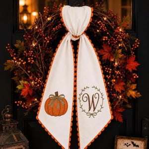 May include: A decorative wreath featuring autumn foliage, orange berries, and warm string lights. A white fabric sash with orange scalloped trim hangs from the center, embroidered with an orange pumpkin on one side and a letter W in a wreath.
