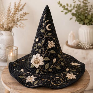 May include: A black quilted pointed hat featuring intricate embroidery of white flowers, green leaves, and a crescent moon. The fabric is detailed with gold stitching and small star motifs that extend from the brim up to the peak of the hat.