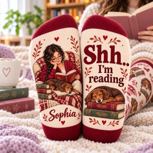 May include: A pair of cream-colored socks with maroon cuffs and toes. One sock features a graphic of a person reading with a dog, and the name Sophia. The other sock displays the text Shh.. I'm reading above a sleeping dog on a stack of books.