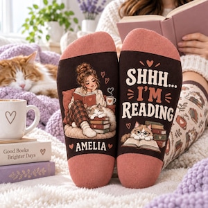 May include: A pair of brown and dusty pink socks featuring a graphic of a person reading with a cat on a stack of books, the text "SHHH... I'M READING" and the name "AMELIA" printed in white, with small heart accents throughout the design.