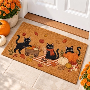 May include: A rectangular tan coir doormat featuring a fall-themed illustration of three black cats, pumpkins, a wooden wagon, a checkered picnic blanket, autumn leaves, acorns, and mushrooms, all arranged in a festive seasonal scene.