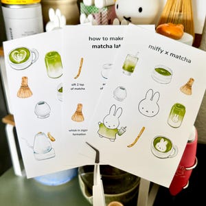 Op de afbeelding: Drie witte kaarten met matcha latte illustraties en tekst. E&eacute;n kaart leest "how to make matcha lat" en een andere "miffy x matcha". De kaarten tonen een garde, een konijn en een kopje matcha. Een pincet staat op de voorgrond.