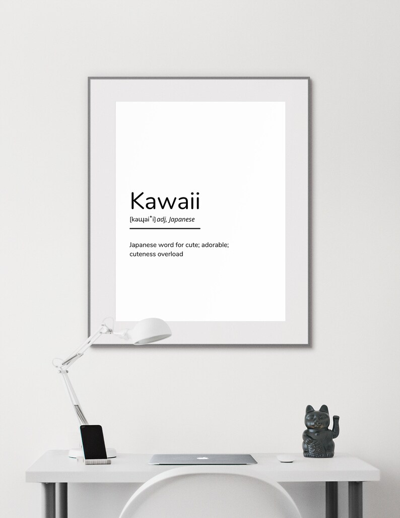May include: A black and white printable wall art print with the word "Kawaii" in bold, black text. The word is defined as a Japanese word for cute, adorable, and cuteness overload. The print is framed in a black frame and is sitting on a white desk with a lamp and a black cat figurine.