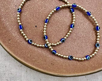 May include: Two gold bracelets with blue evil eye charms. The bracelets are layered on top of each other.