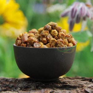 May include: A brown ceramic bowl filled with bee pollen granules. The granules are a light brown color and have a slightly sticky texture.