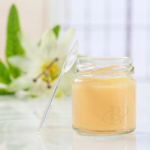 Pode incluir: Um pequeno frasco de vidro transparente preenchido com uma substância amarelo claro, possivelmente mel. Uma colher de plástico transparente está encostada no frasco. Uma flor branca desfocada e folhas verdes ao fundo.