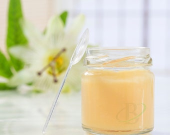 May include: A small, clear glass jar filled with a light yellow substance, possibly honey or a similar product. A clear plastic spoon rests against the jar. A blurred white flower and green leaves are in the background.