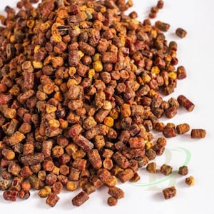 May include: A pile of bee pollen granules featuring a variety of textures and shapes in shades of deep reddish-brown, golden yellow, and burnt orange. The small, cylindrical pellets are clustered together, creating a dense, granular mound.