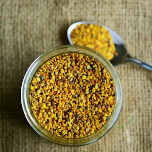 May include: A glass jar filled with yellow and brown bee pollen, with a spoon full of bee pollen in the background.