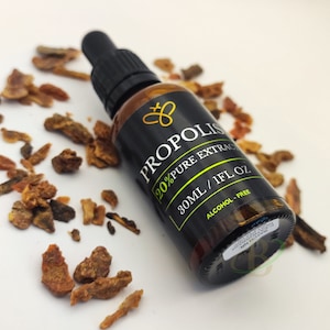 May include: A dark brown glass bottle with a black label that says "Propolis 20% Pure Extract 30ml / 1fl oz Alcohol-Free". The bottle has a dropper and is surrounded by small pieces of brown and orange propolis.